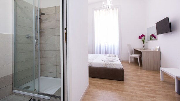 A modern hotel room with a glass-enclosed shower, a bed with a white duvet, a desk with a chair, and a flat-screen TV mounted on the wall. - El Dorado Vatican (Rome)
