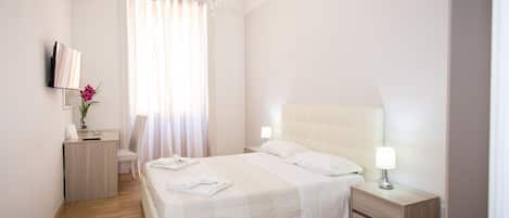 A modern hotel room with a large bed, bedside tables, a desk, a chair, and a TV on the wall.