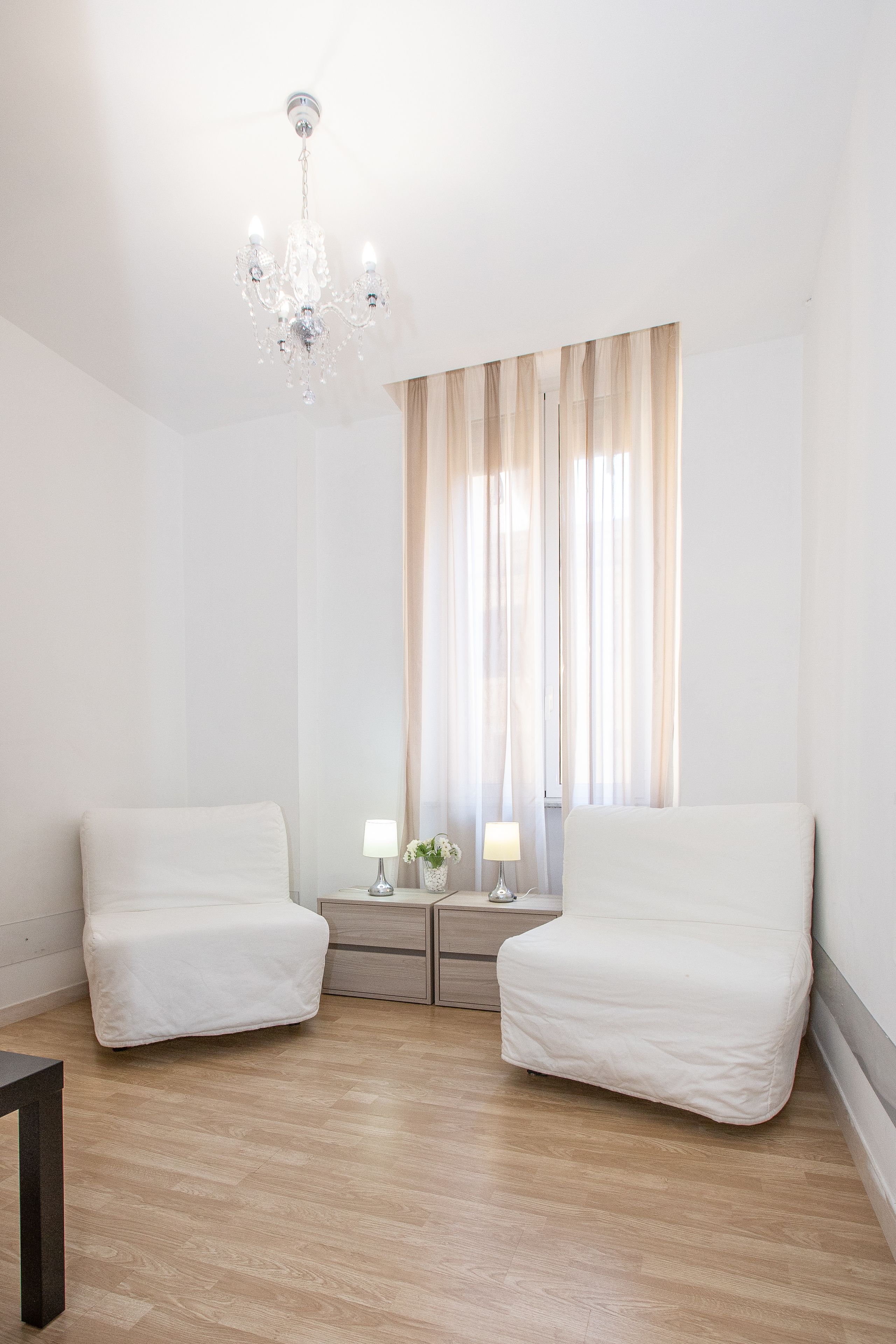 A room with two white chairs, a small table, a nightstand with lamps, and a chandelier.