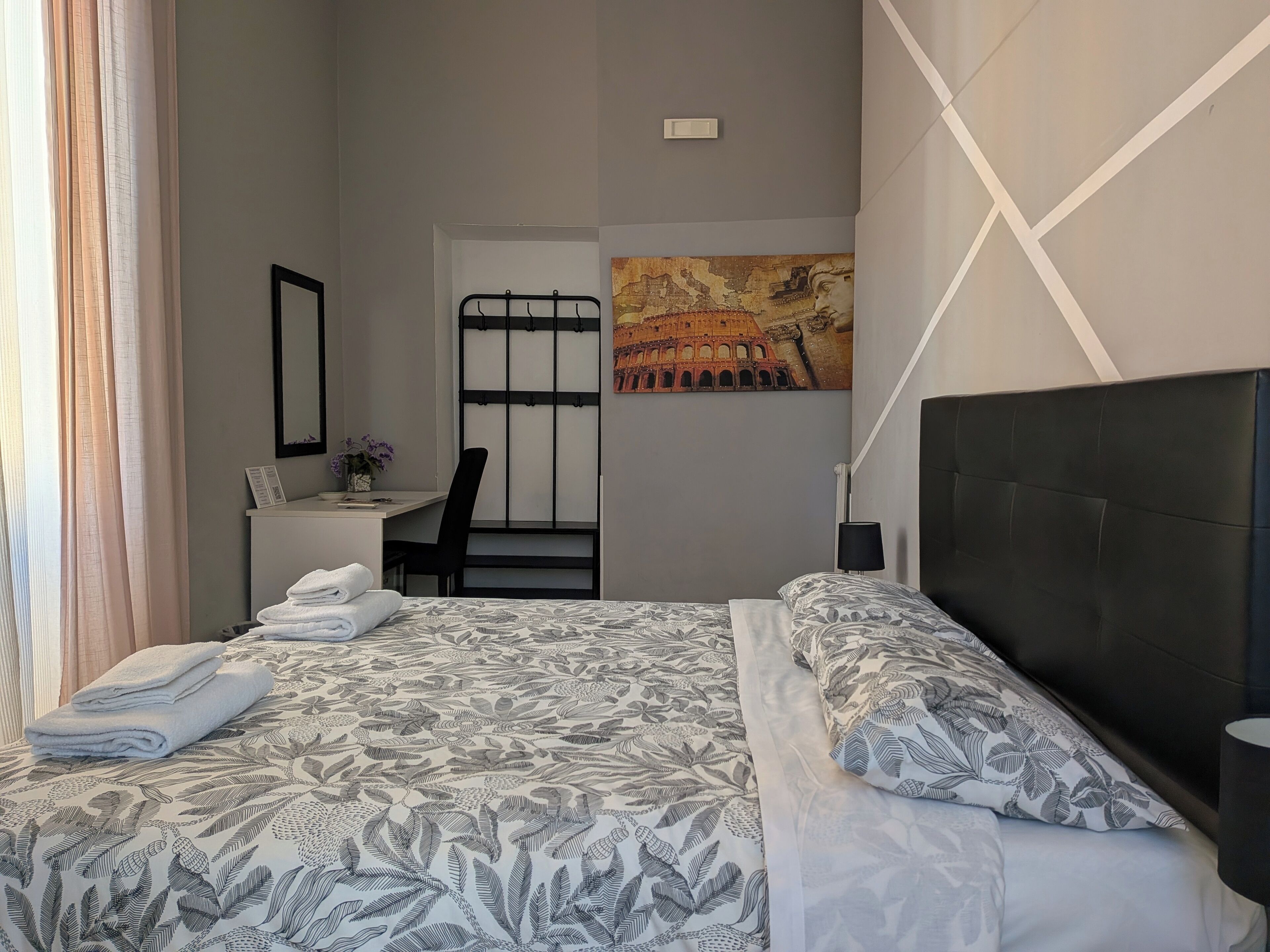 A hotel room with a bed, a desk, a chair, a painting of the Colosseum, and a window with curtains.