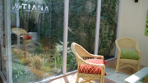 Una habitación con amplios ventanales de vidrio, sillas de mimbre con cojines y un jardín exterior visible.