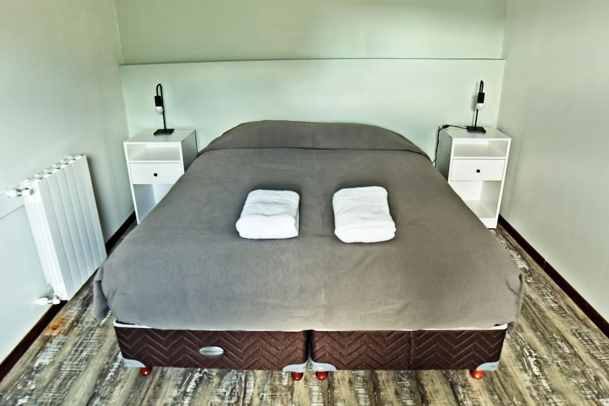 Un lit bien fait, avec deux serviettes pliées, une table de chevet blanche avec une lampe et un radiateur au mur.