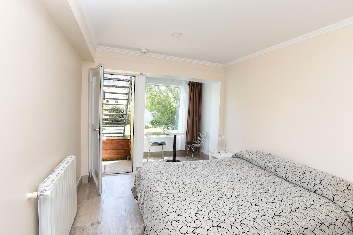 Une chambre avec un lit, un radiateur, une fenêtre et une porte ouverte donnant sur un espace extérieur équipé d’une table et de chaises.