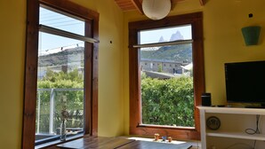 Ein Zimmer mit Holzbalken, großen Fenstern mit Blick auf die Berge, einem weißen Regal mit Fernseher und einer Pendelleuchte.