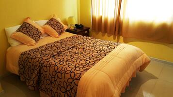 Una cama bien hecha con una colcha estampada y almohadas a juego, una mesita de noche con lámpara y teléfono, y una ventana con cortinas.