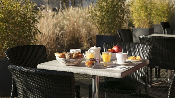 Un área de comedor al aire libre con sillas de mimbre y una mesa preparada con alimentos para el desayuno, incluyendo pan, jugo y un bol de fruta.