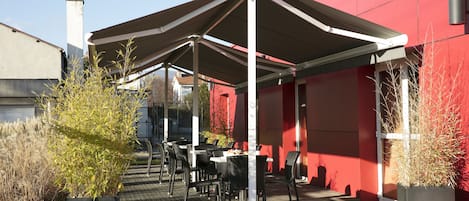Un área de estar al aire libre cubierta, con mesas y sillas, rodeada de plantas en macetas y un edificio rojo.