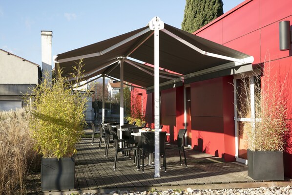 Un área de estar al aire libre cubierta, con mesas y sillas, rodeada de plantas en macetas y un edificio rojo.