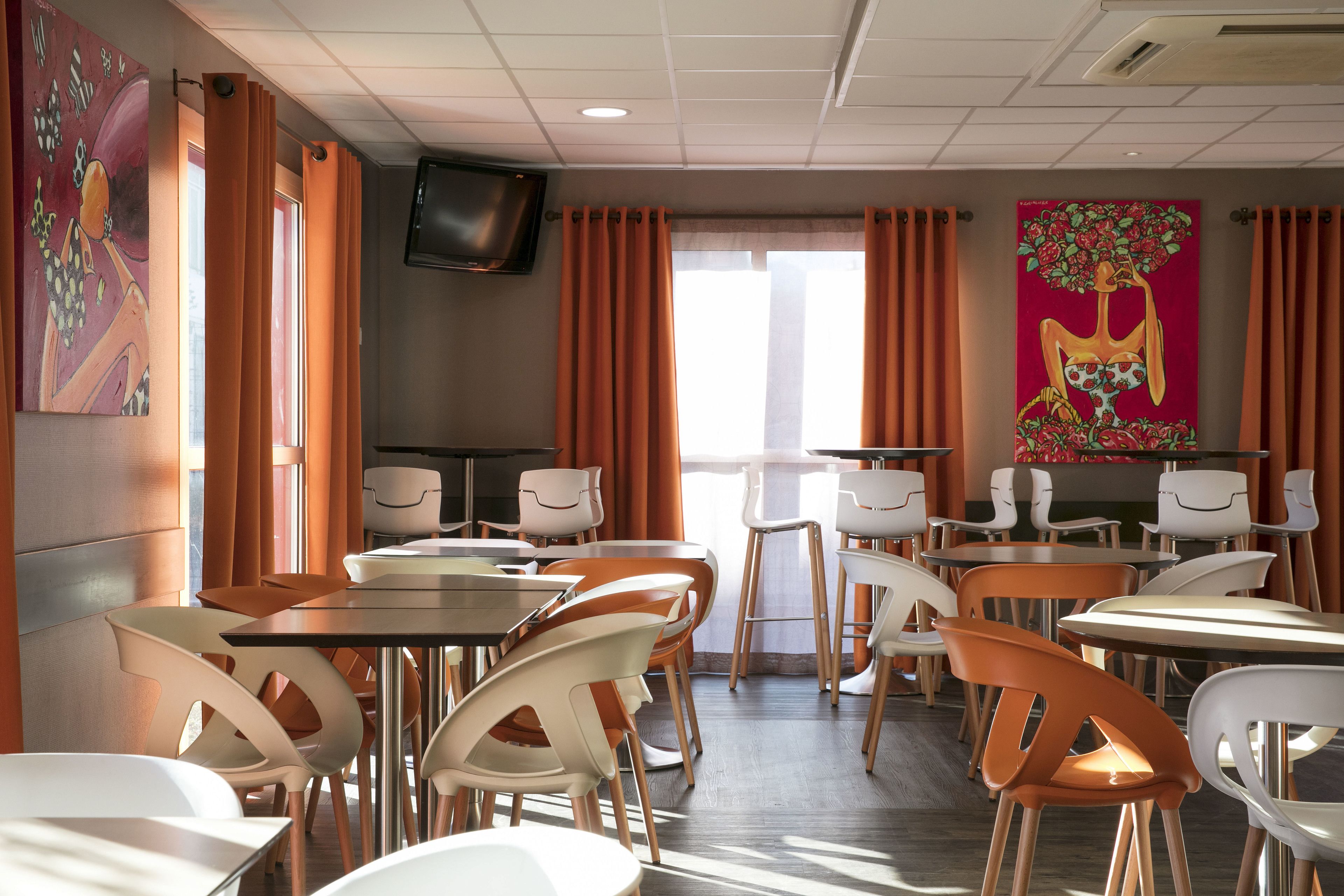 A dining area with tables and chairs arranged in a casual layout, a flat-screen TV mounted on the wall, and a window with curtains.