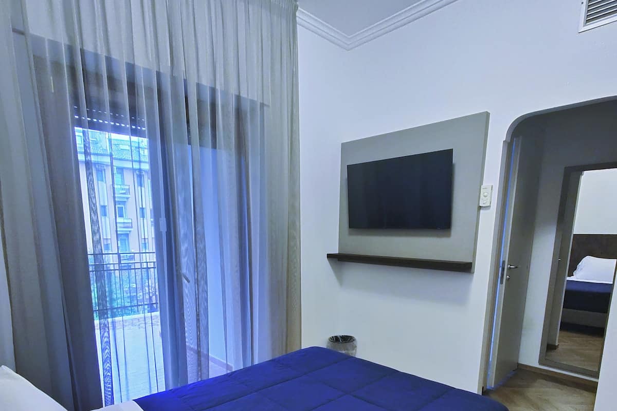 Habitación de hotel con cama, televisión montada en la pared, ventana con cortinas y puerta que da a otra habitación.