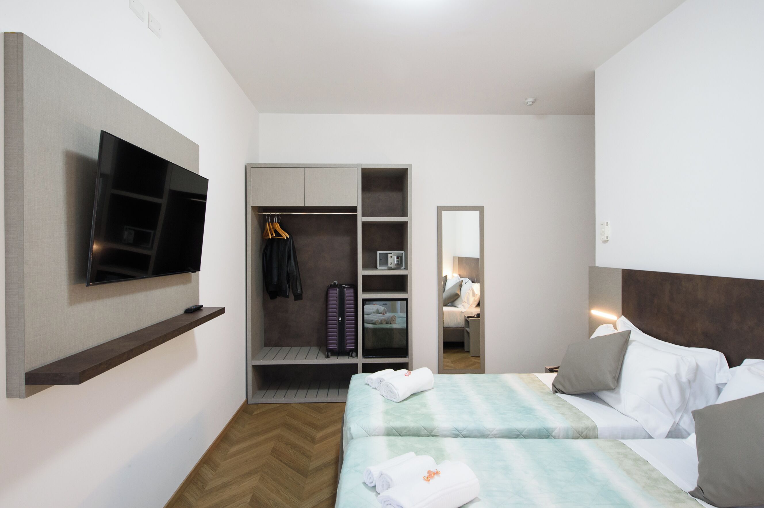 A hotel room with a TV mounted on the wall, a wardrobe with hanging clothes, a bed with pillows, and a bedside table with a lamp.