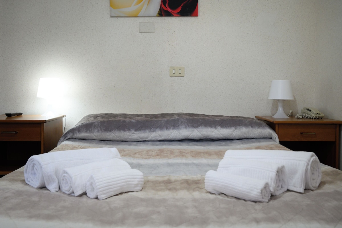 Una cama bien hecha con dos toallas enrolladas, una lámpara y un teléfono sobre una mesita de noche.