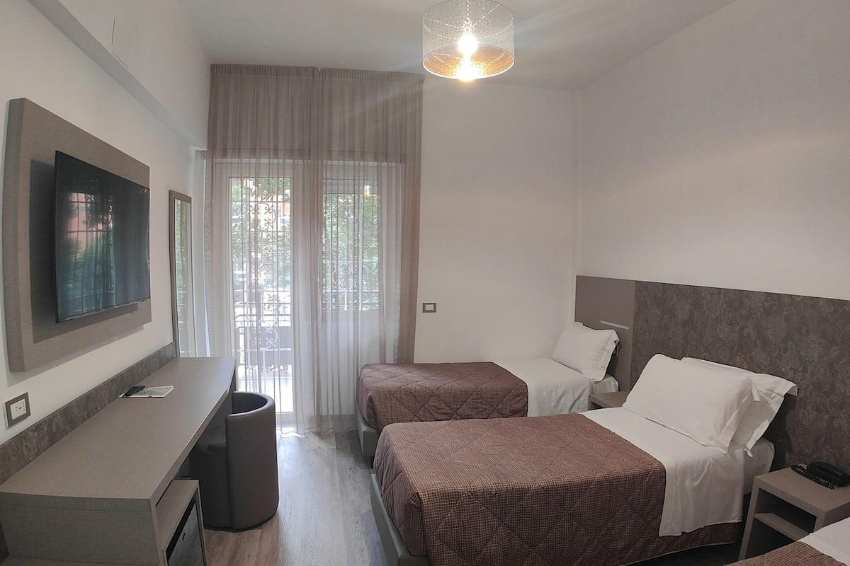 Habitación de hotel con dos camas, un escritorio, televisión y un ventanal con cortinas.