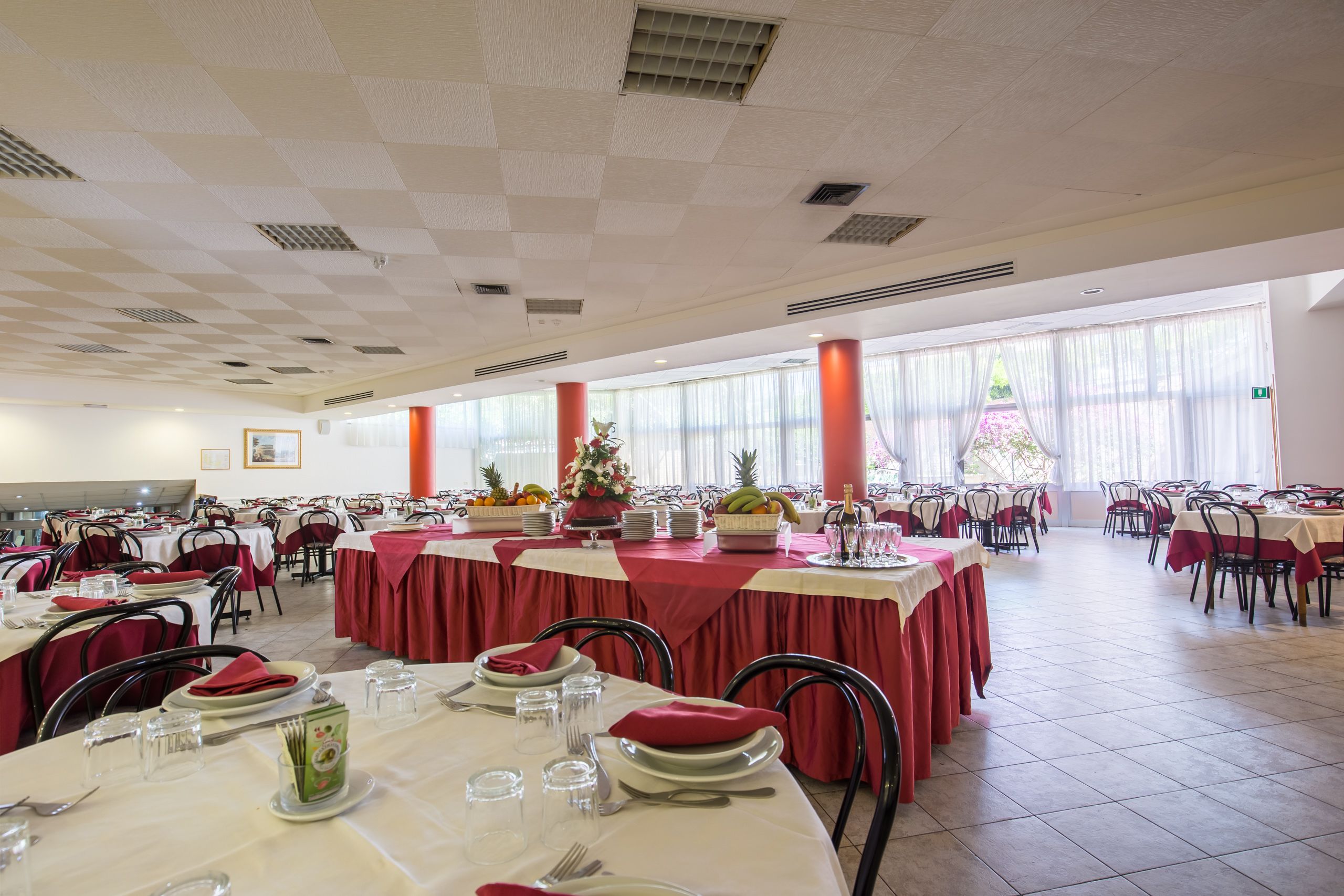 A large dining hall with multiple tables set for service, featuring red tablecloths and white plates.