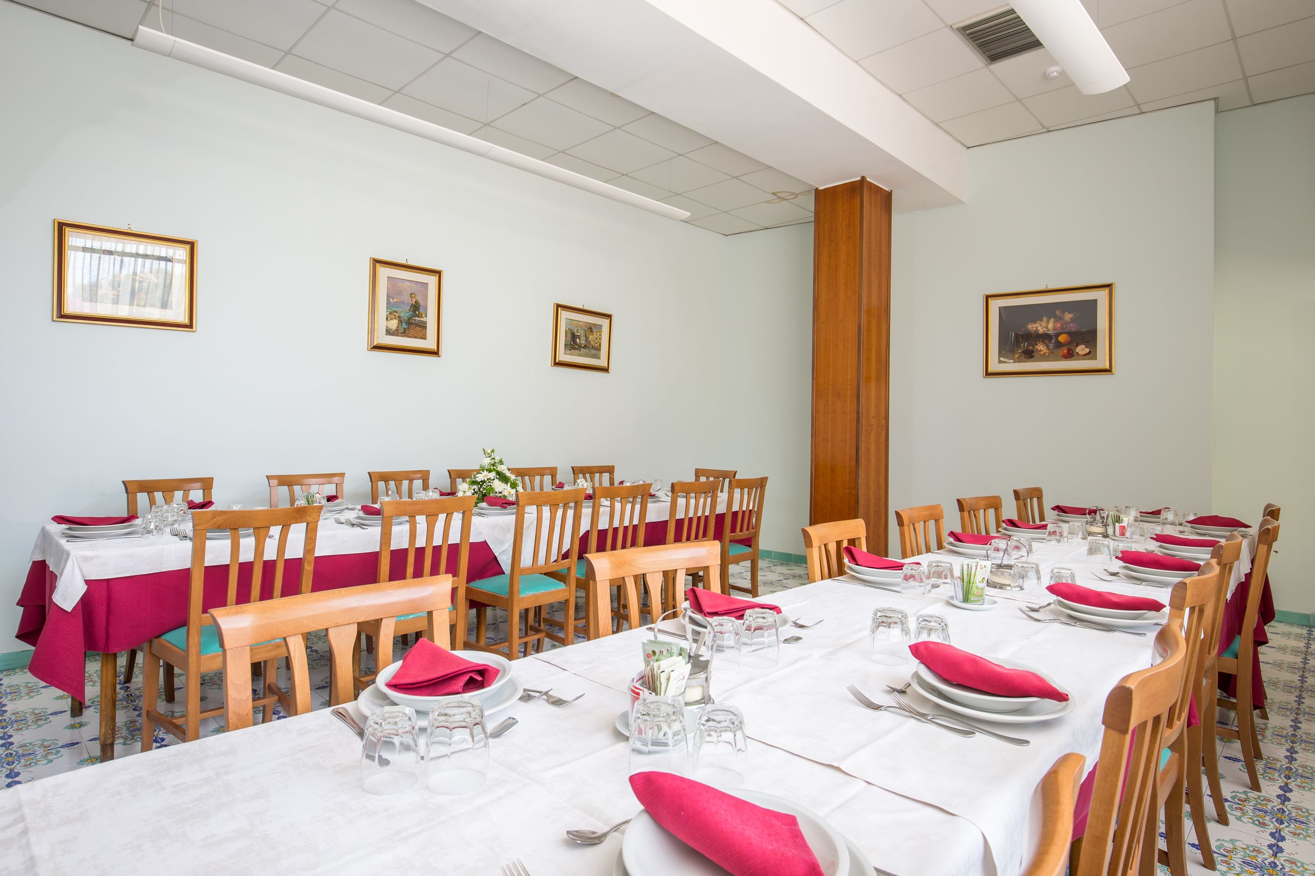 Un comedor con varias mesas dispuestas para una comida, sillas de madera y cuadros en las paredes.
