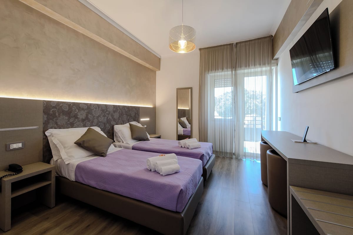 Habitación de hotel con dos camas, un escritorio, un televisor y un ventanal con cortinas.