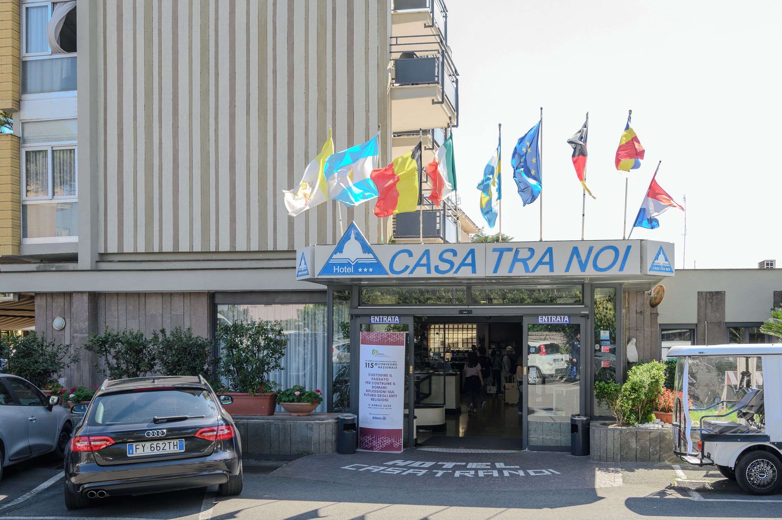 La entrada del Hotel CASA TRA NOI con varias banderas, un área de estacionamiento y servicio de transporte.