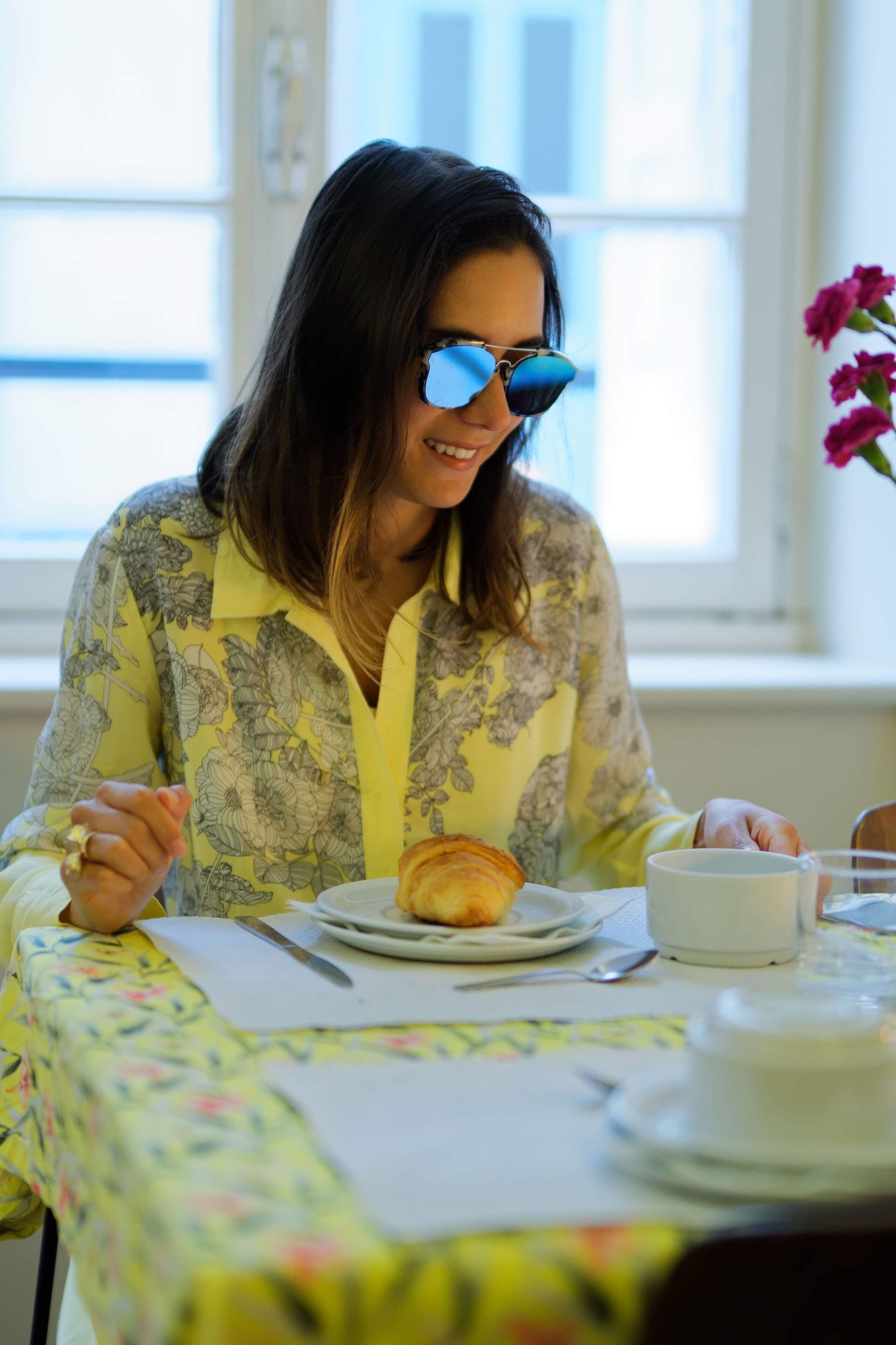 A person sitting at a table with a croissant and a cup, in a room with a window and a vase of flowers.