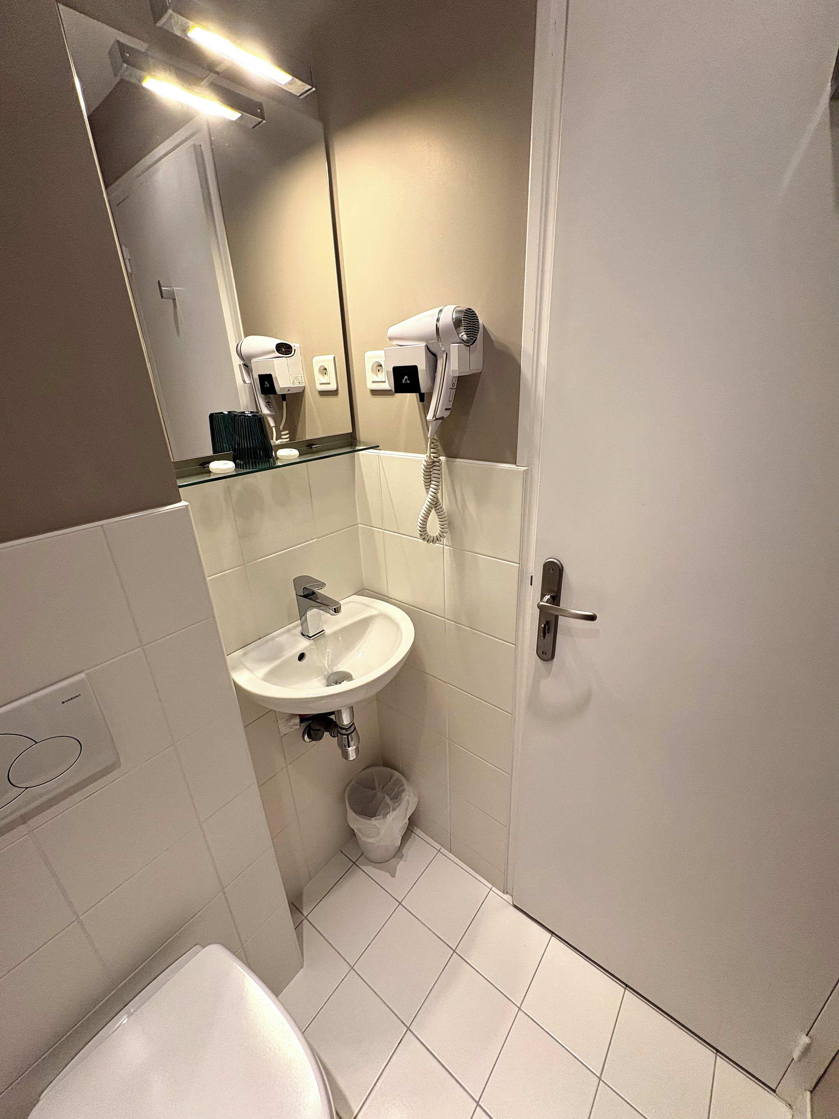 Een kleine, schone badkamer met een witte wasbak, een spiegel, een haardroger en een prullenbak.