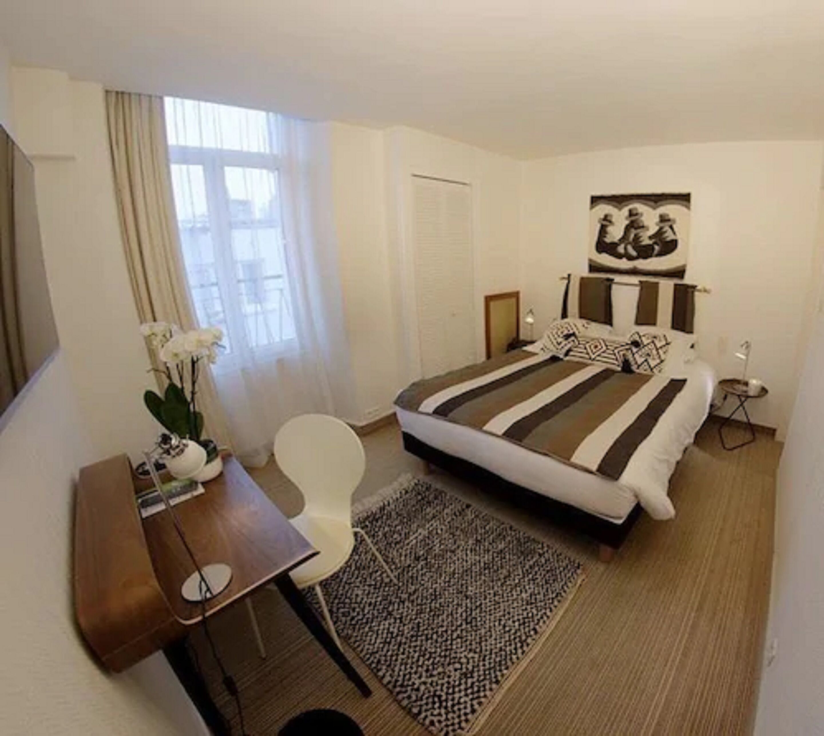 Una camera d'albergo con un letto, una scrivania, una sedia, un tavolino, una lampada, un quadro e una finestra con tende.