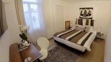 Una camera d'albergo con un letto, una scrivania, una sedia, un tavolino, una lampada, un quadro e una finestra con tende.