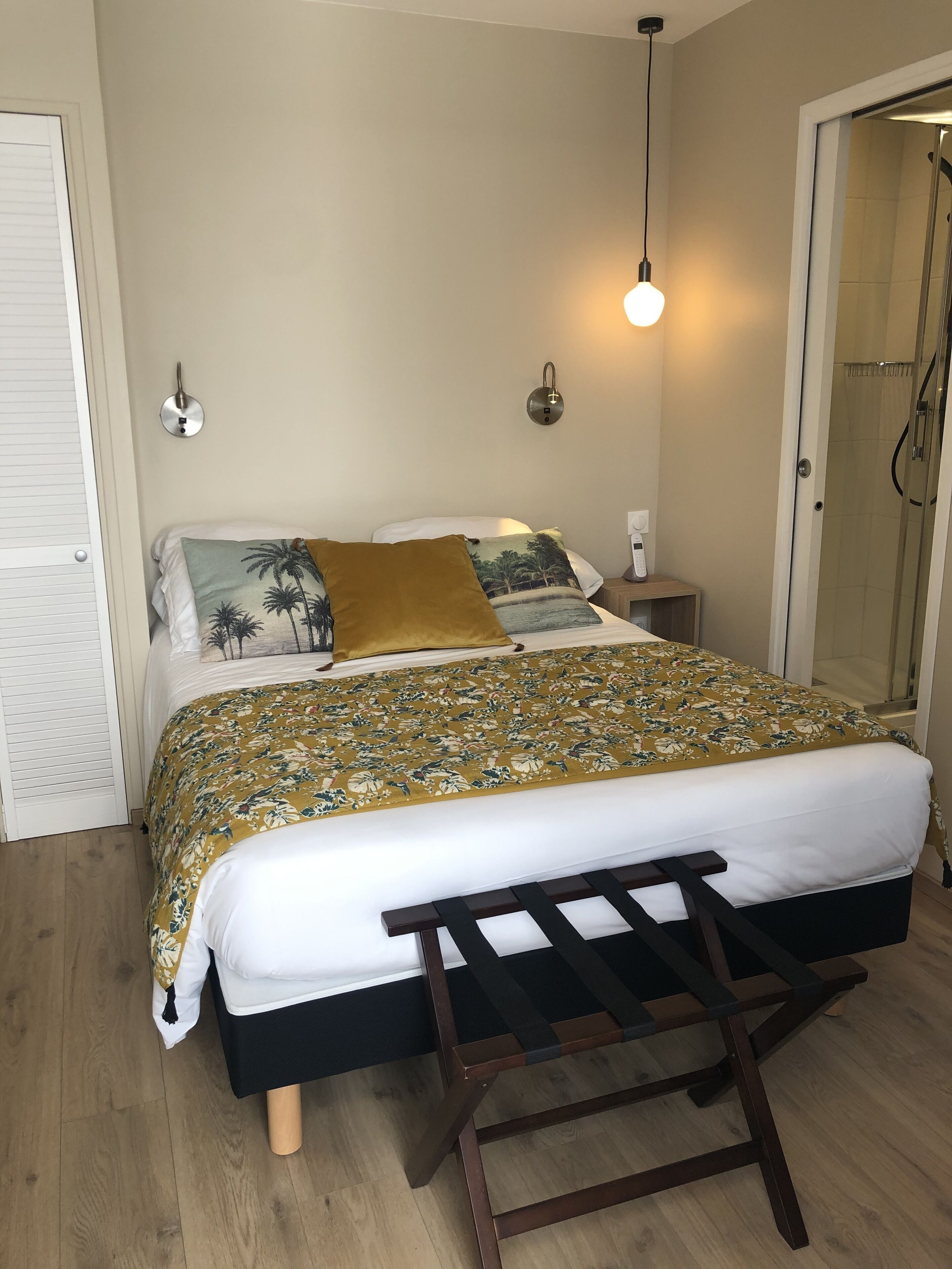 A bedroom with a bed, a bedside table, a nightstand, a wall-mounted light, and a door leading to a shower area.