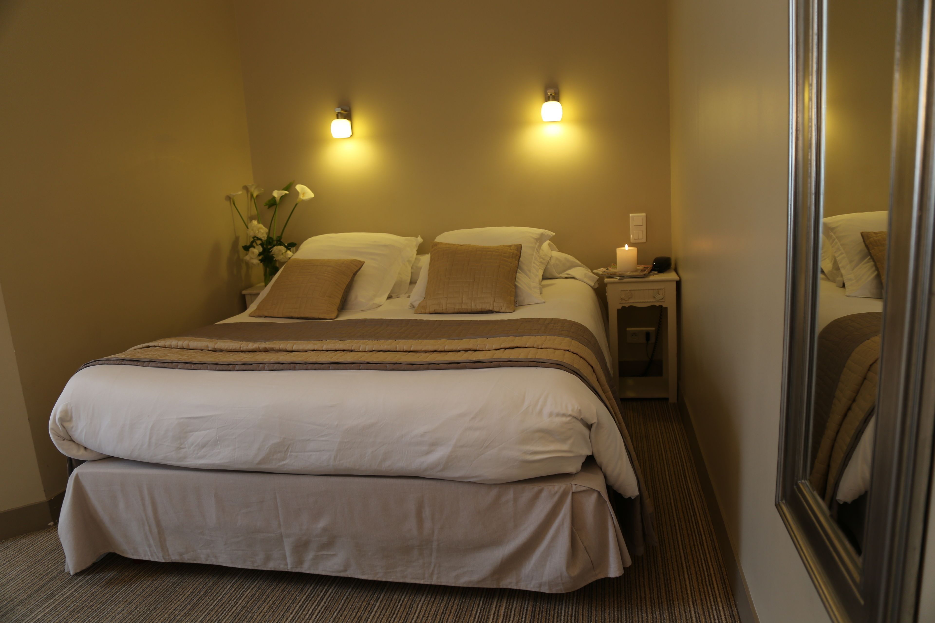 Habitación de hotel con una cama doble, dos mesitas de noche, un tocador, un espejo y un florero con flores.