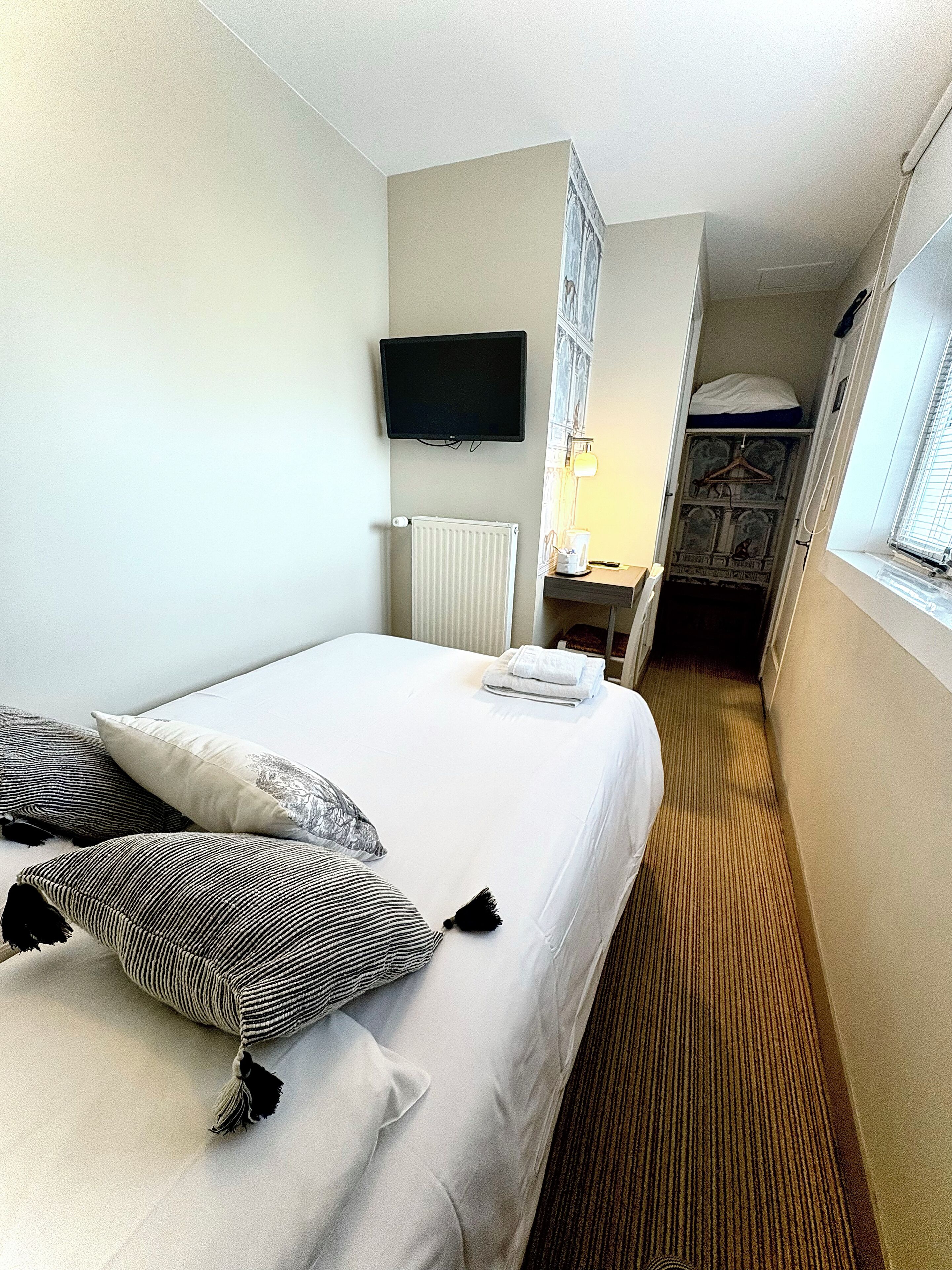 Une chambre d’hôtel avec un lit, des oreillers, une télévision, un radiateur, un bureau avec une lampe et une fenêtre.