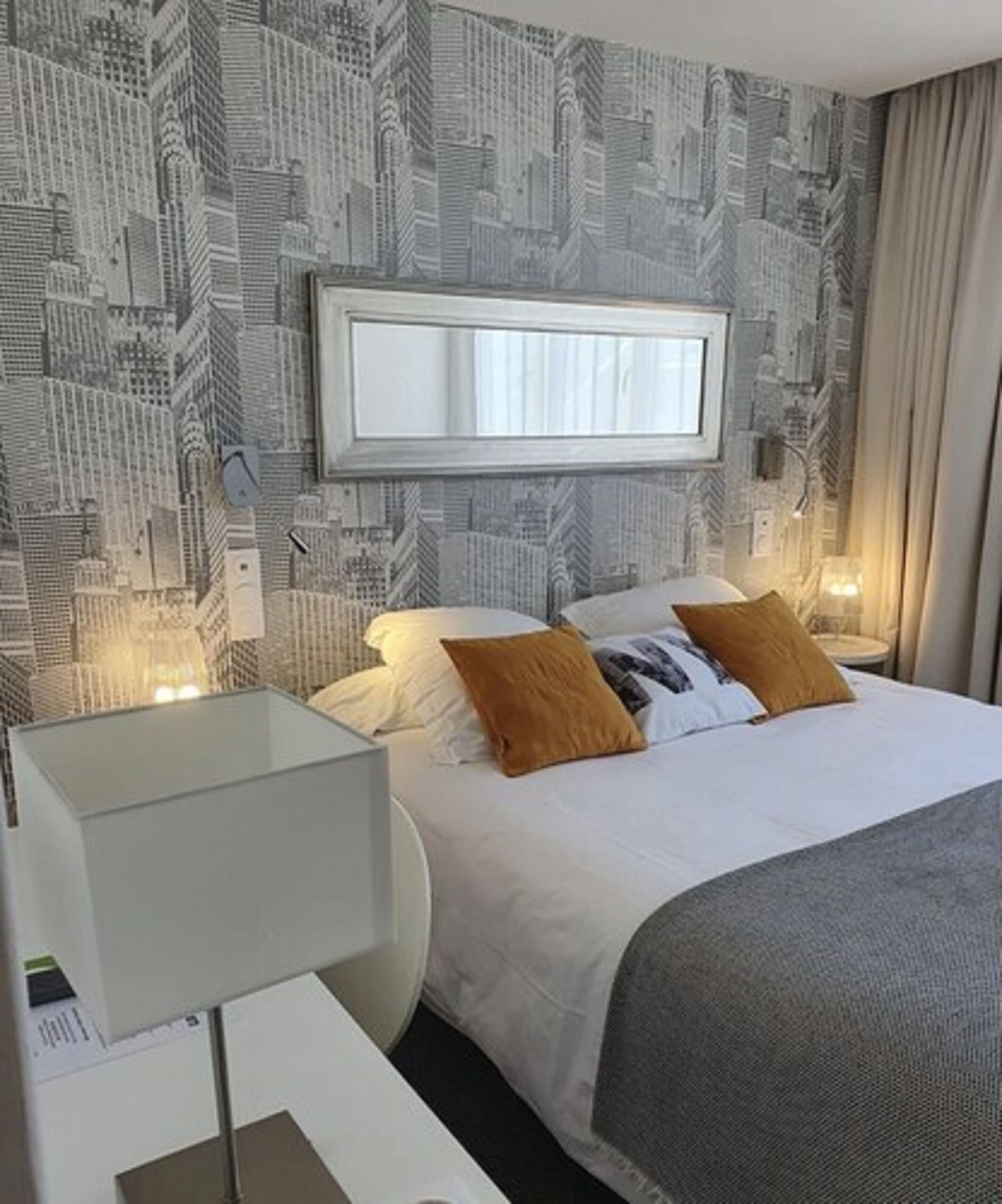 A hotel room with a bed, a bedside table, a lamp, and a wall with a cityscape wallpaper.