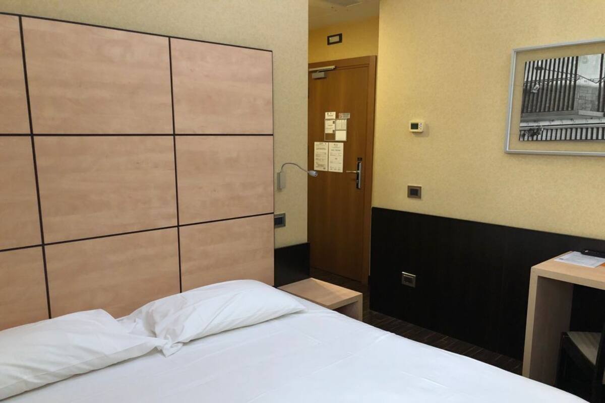 A hotel room with a bed, a wooden headboard, a small bedside table, a nightstand, a desk, and a framed picture on the wall.