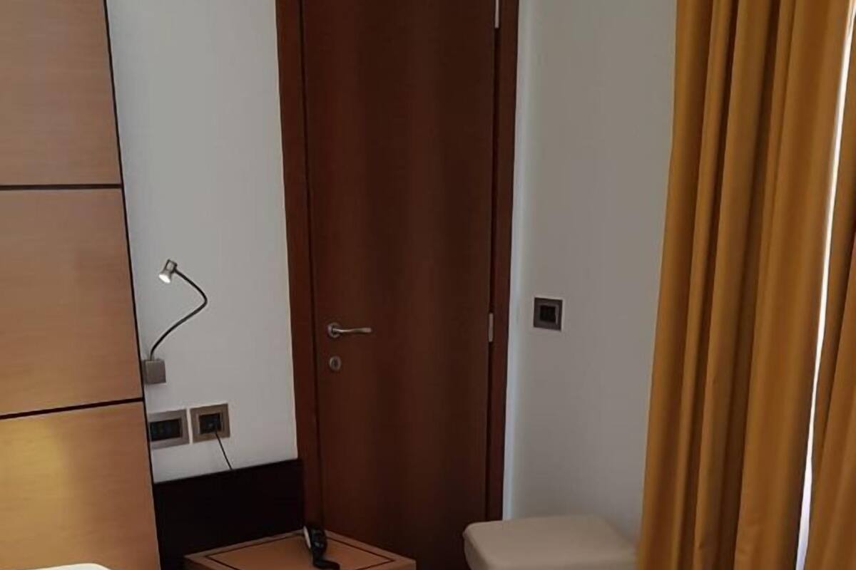 A hotel room with a single bed, a bedside table, a nightstand, a wooden wardrobe, a toilet, and a curtain.