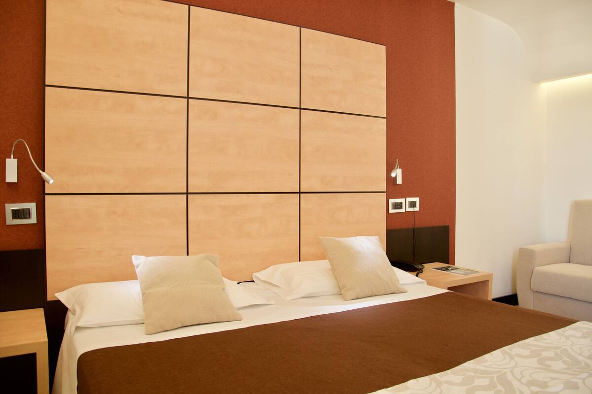 A hotel room with a large bed, a bedside table, a chair, and a wall-mounted TV.