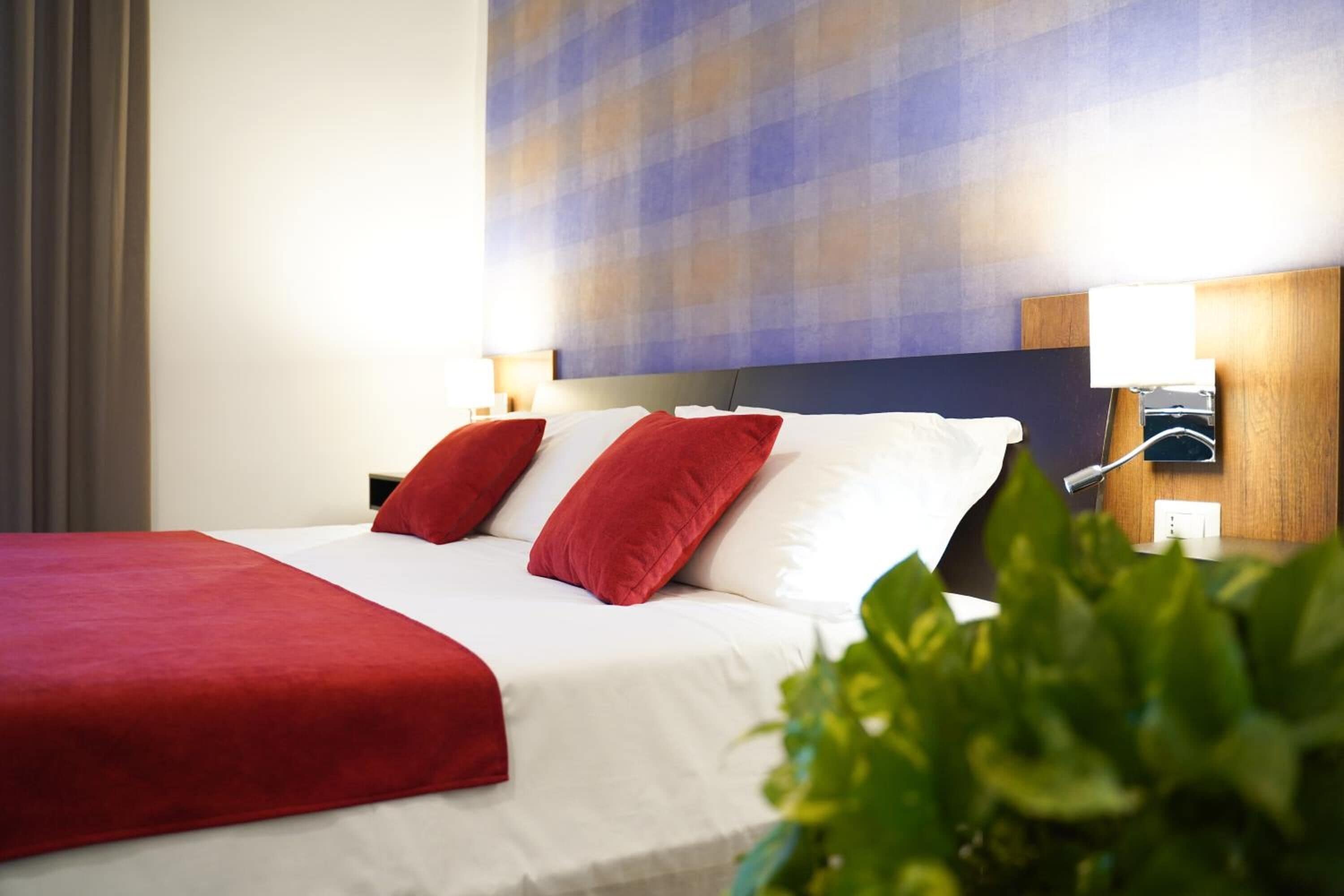 Une chambre d’hôtel avec un lit, des oreillers rouges, un couvre-lit blanc, un mur bleu et orange, et une plante verte.