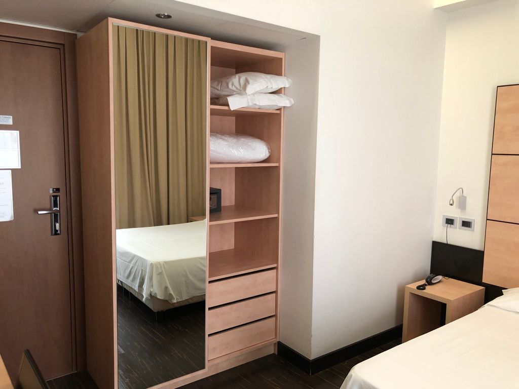 Une chambre d’hôtel comprenant un lit, une armoire en bois avec un rideau, une table de chevet avec un téléphone et une lampe fixée au mur.