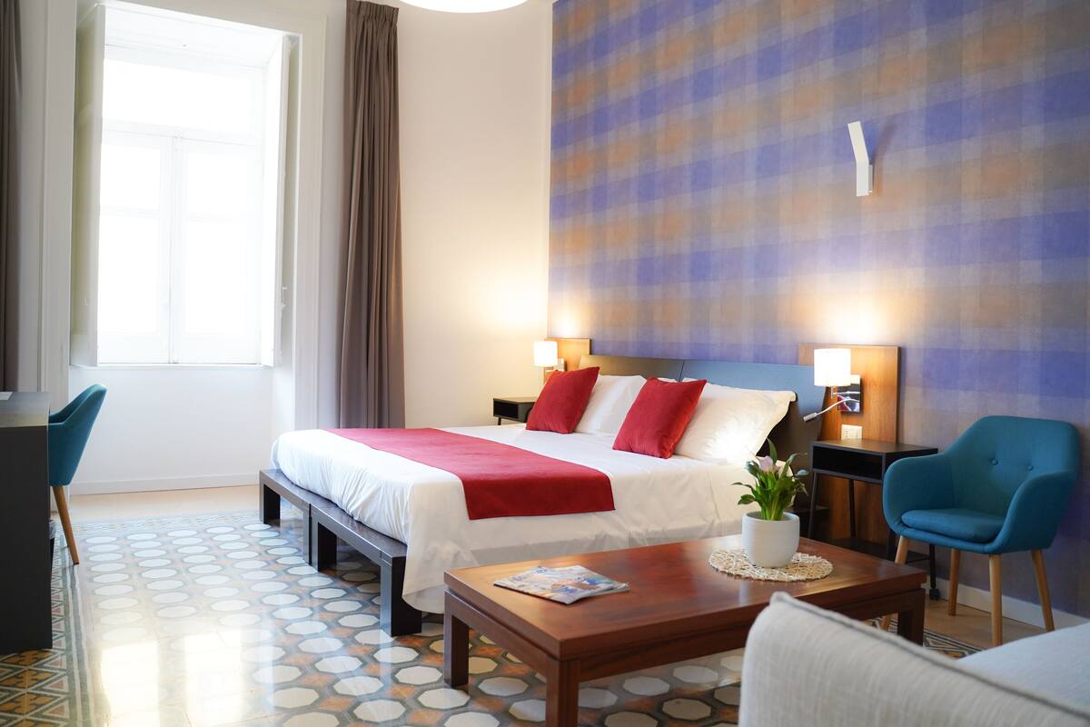 A hotel room with a bed, bedside tables, a desk, a chair, a coffee table, and a potted plant.