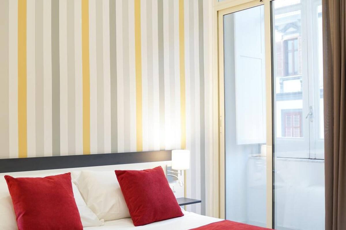 A hotel room with a bed, red and white pillows, a red blanket, a striped wall, and a sliding glass door to the balcony.