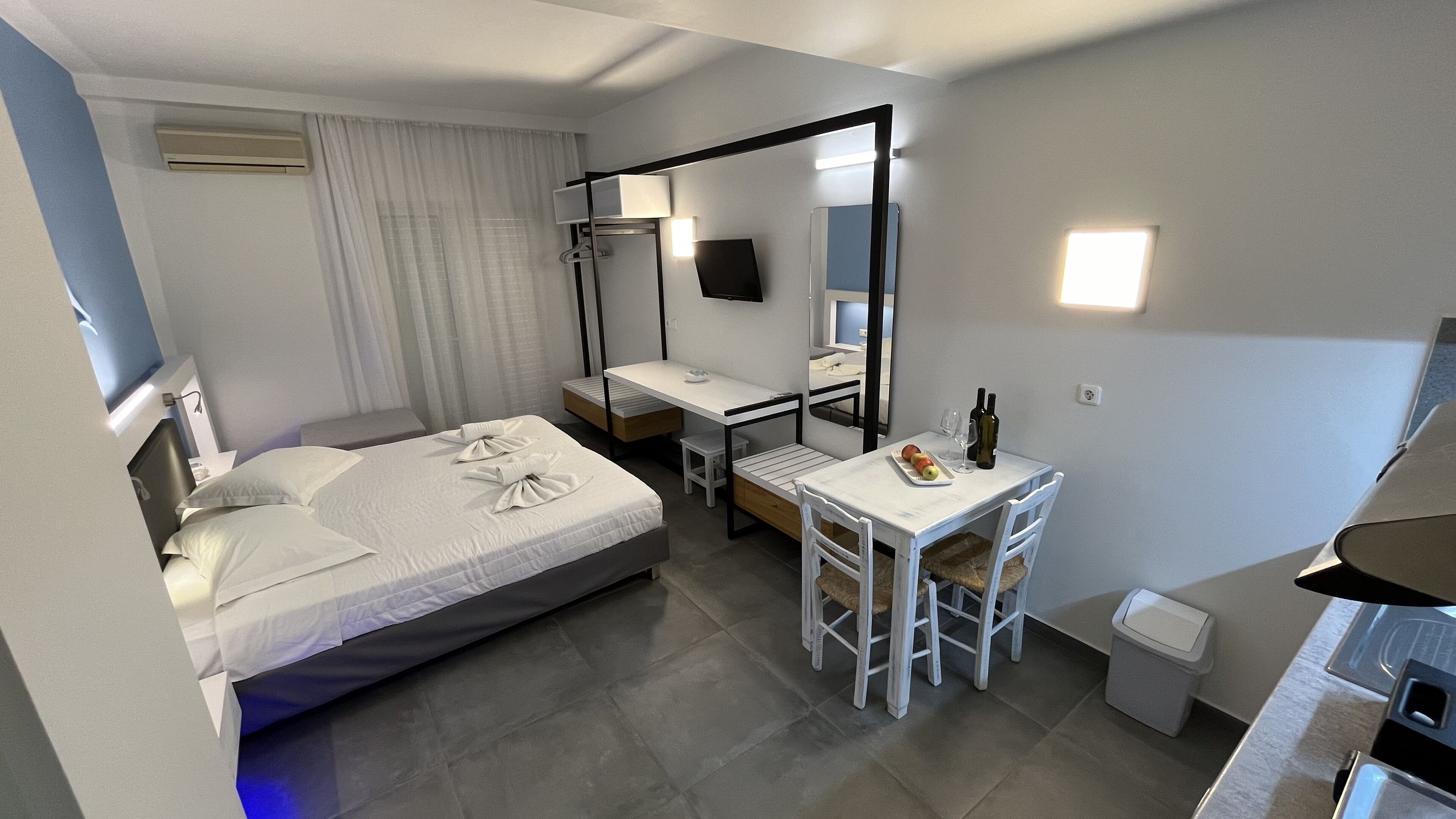 A hotel room with a bed, a desk, a chair, a mirror, a TV, and a small kitchenette.
