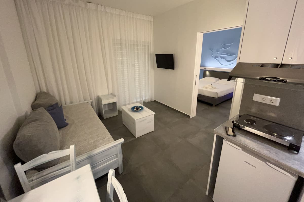 A compact hotel room with a bed, a small bedside table, a TV, a kitchenette, and a window with curtains.