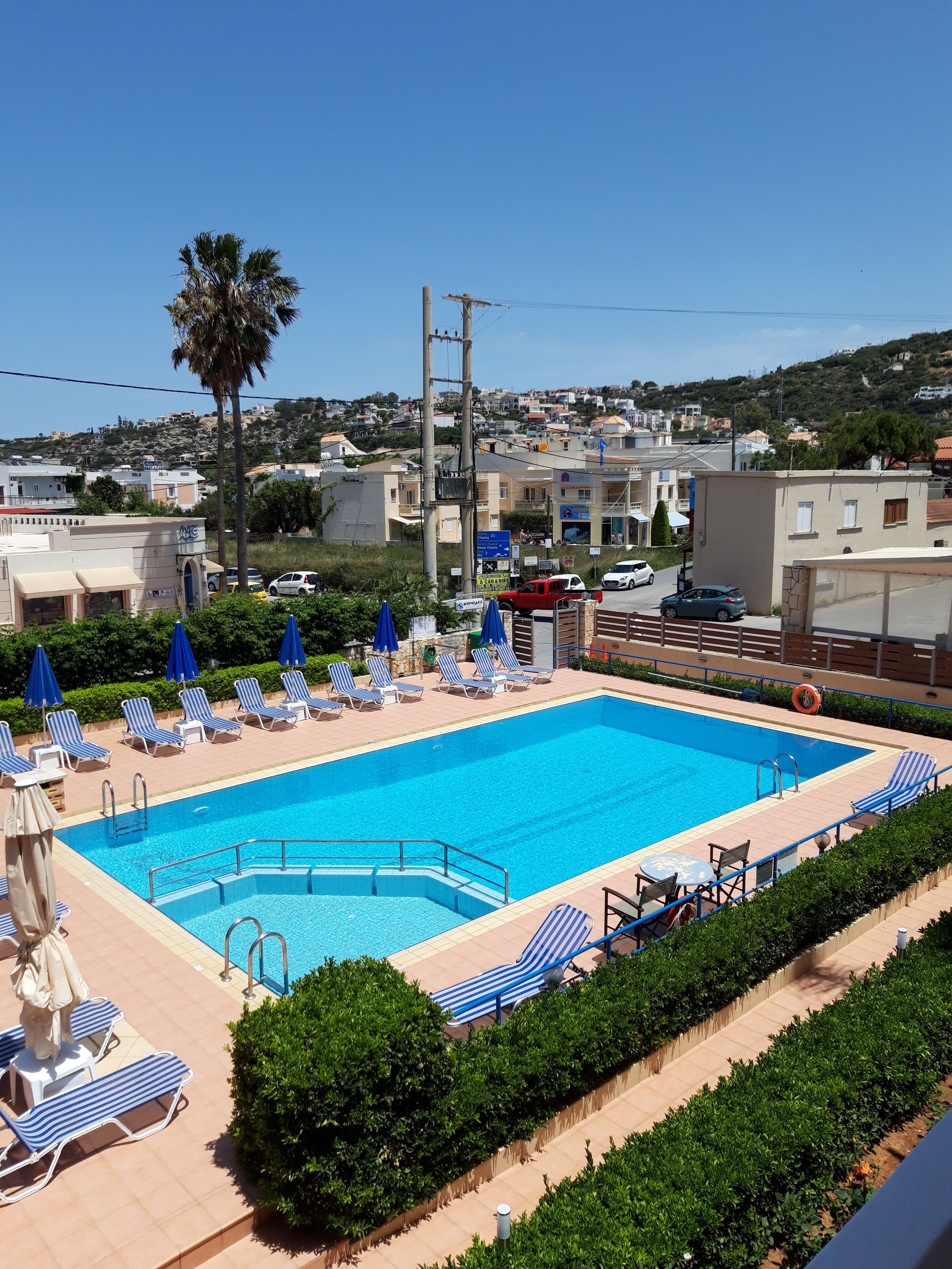 Une piscine entourée de chaises longues et de parasols, avec une végétation luxuriante et des bâtiments en arrière-plan.