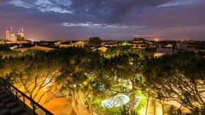 A nighttime view of a cityscape with illuminated buildings and a park area with trees and a circular water feature. - Astromare (Jesolo)