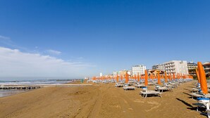 A beach with rows of orange umbrellas and beach chairs, a sandy shore, and buildings in the background. - Astromare (Jesolo)