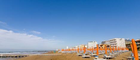 Una playa con hileras de sombrillas y sillas de playa anaranjadas, una orilla arenosa y edificios al fondo.