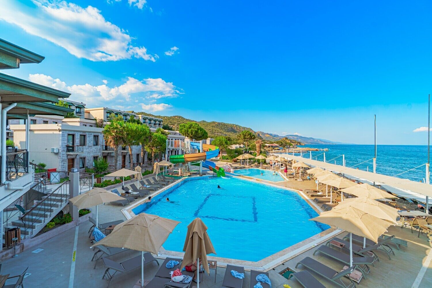 A pool area with a water slide, lounge chairs, umbrellas, and a view of the sea.
