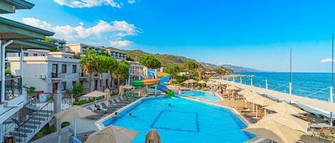 A pool area with a water slide, lounge chairs, umbrellas, and a view of the sea.