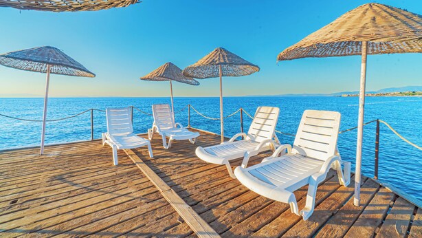 Une terrasse en bois, dotée de chaises longues blanches et de parasols en chaume, surplombe une étendue d’eau.