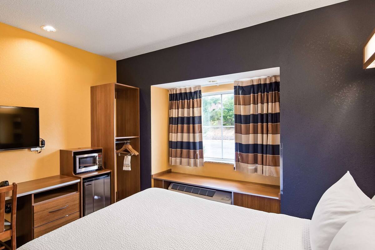 A hotel room with a bed, a desk with a microwave, a TV, and a window with curtains.