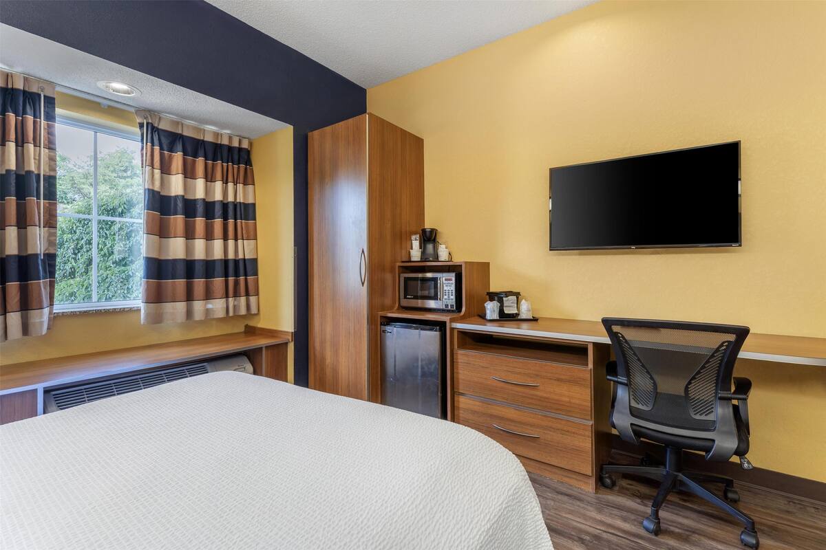 A hotel room with a bed, a desk with a chair, a microwave, a refrigerator, and a flat-screen TV mounted on the wall.