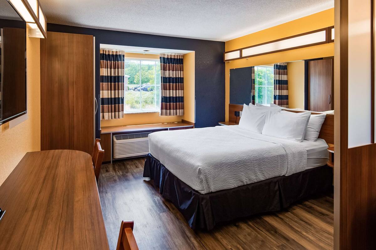 A compact bedroom with a bed, a small table, a window with curtains, and a view of the outdoors.