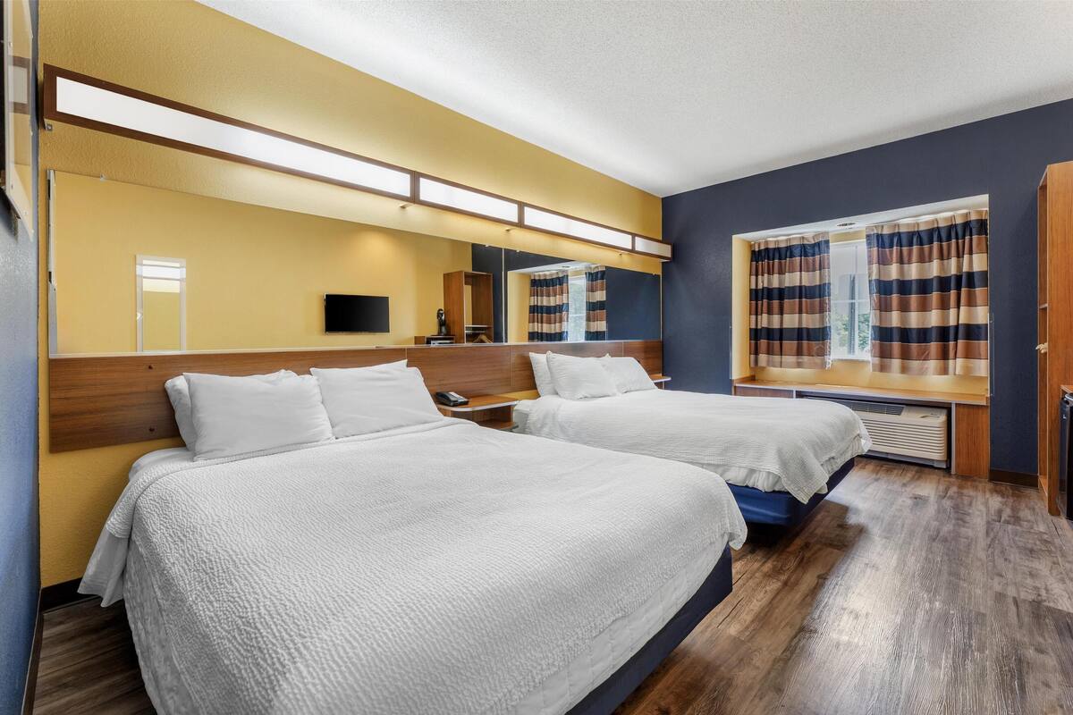 A hotel room with two beds, a large mirror, a flat-screen TV, and a window with striped curtains.