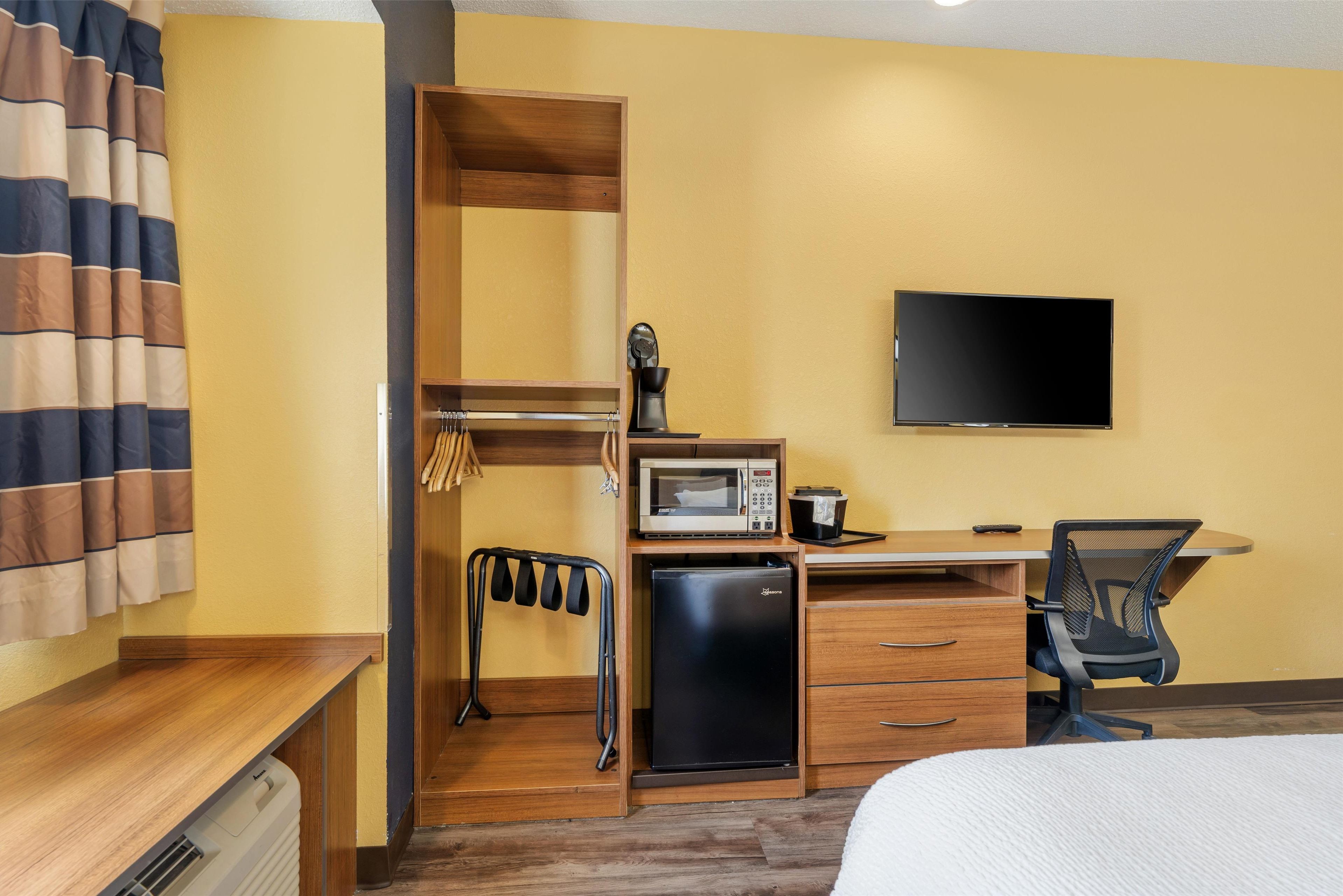 Una habitación de hotel pequeña con una cama, un escritorio con una silla, microondas, refrigerador, televisor y armario.